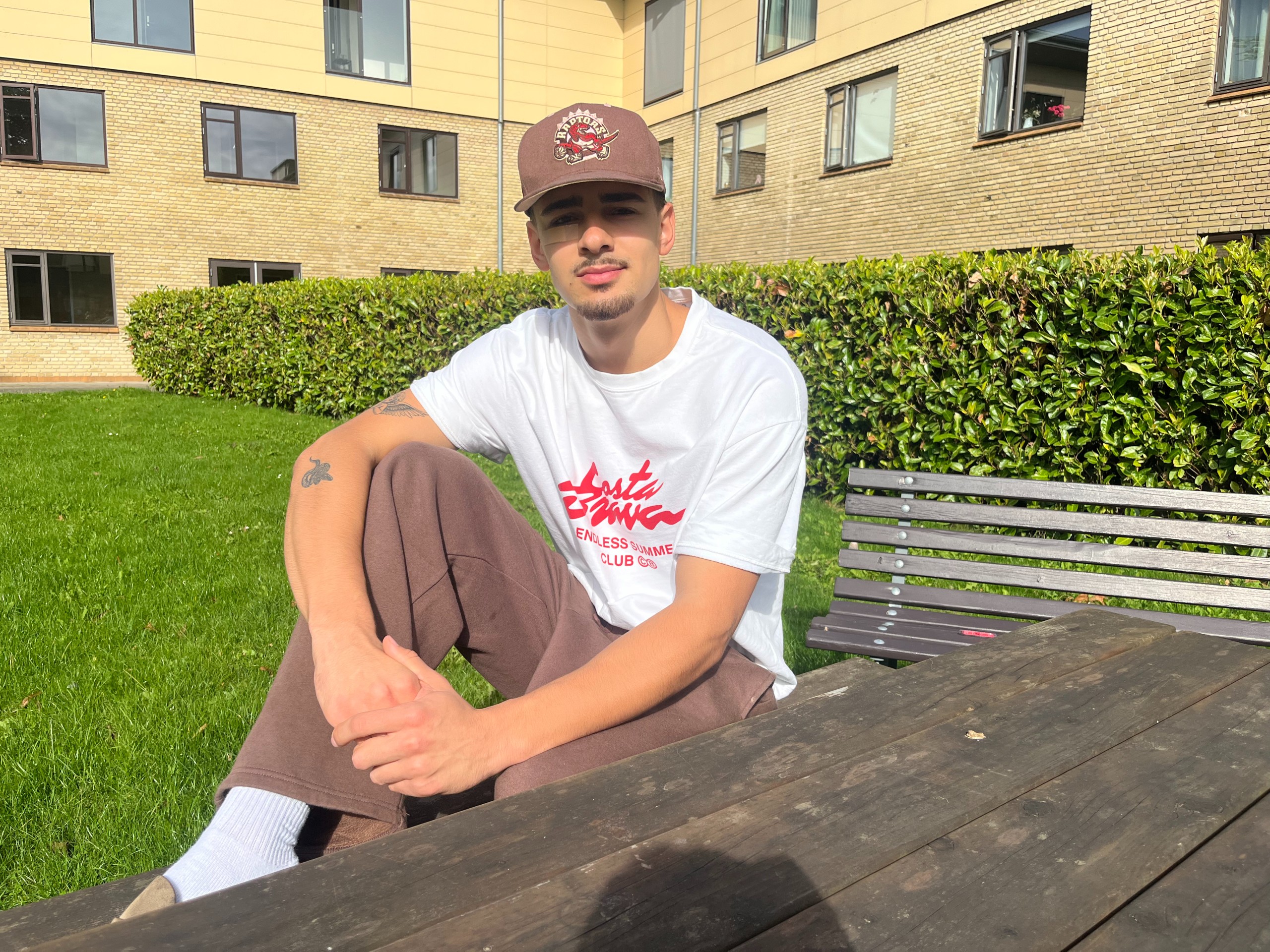 Young man in a cap and white T-shirt sitting at a wooden picnic table outside a brick apartment building