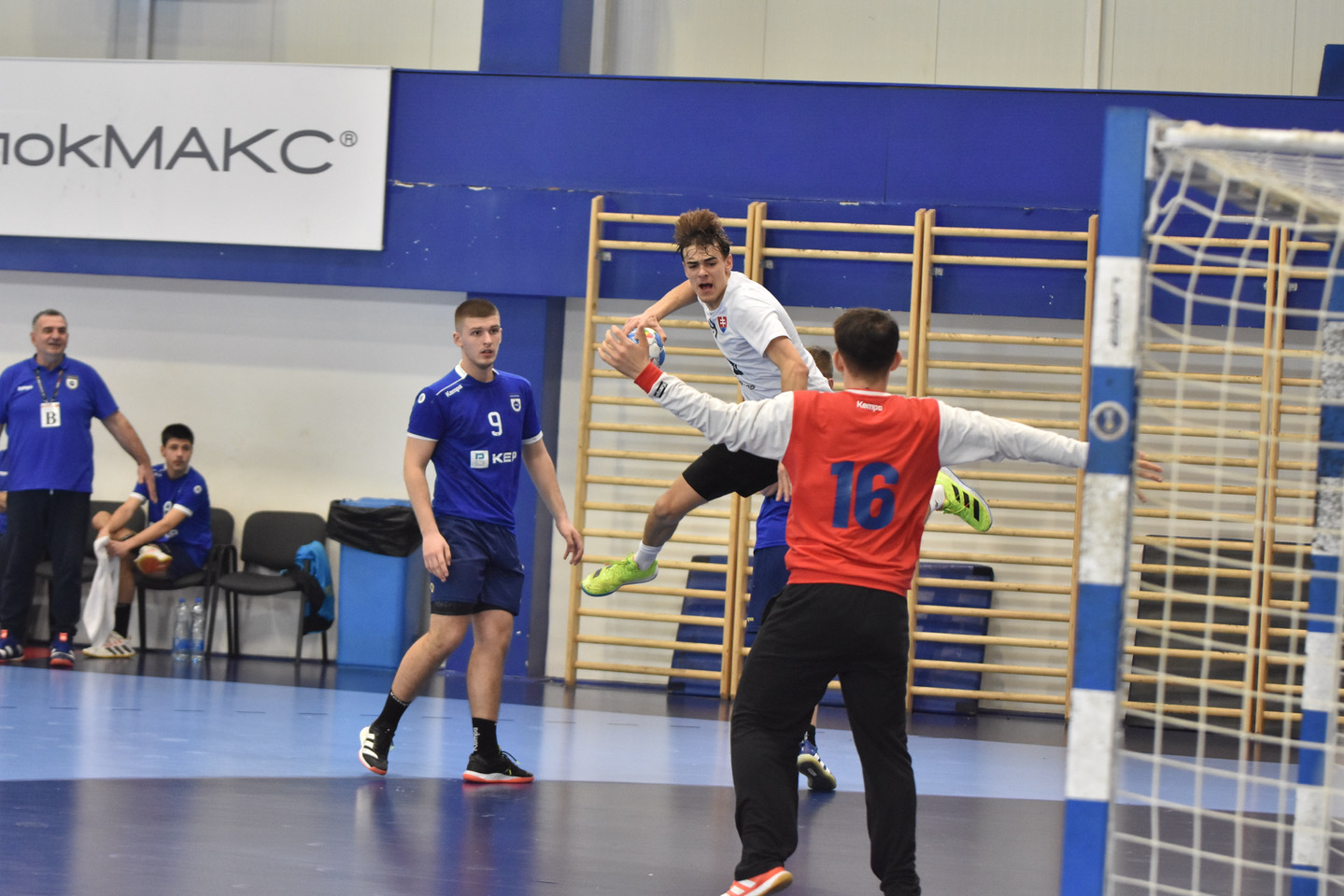 Handball player in a white kit jumping to shoot past a defender in front of the goal during an indoor match