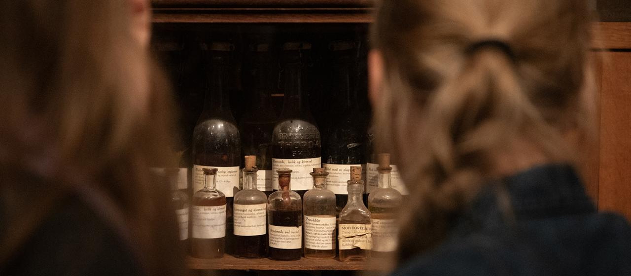 Two people looking into a wooden cabinet of old Danish-labeled glass apothecary bottles and vials, some dark green and amber