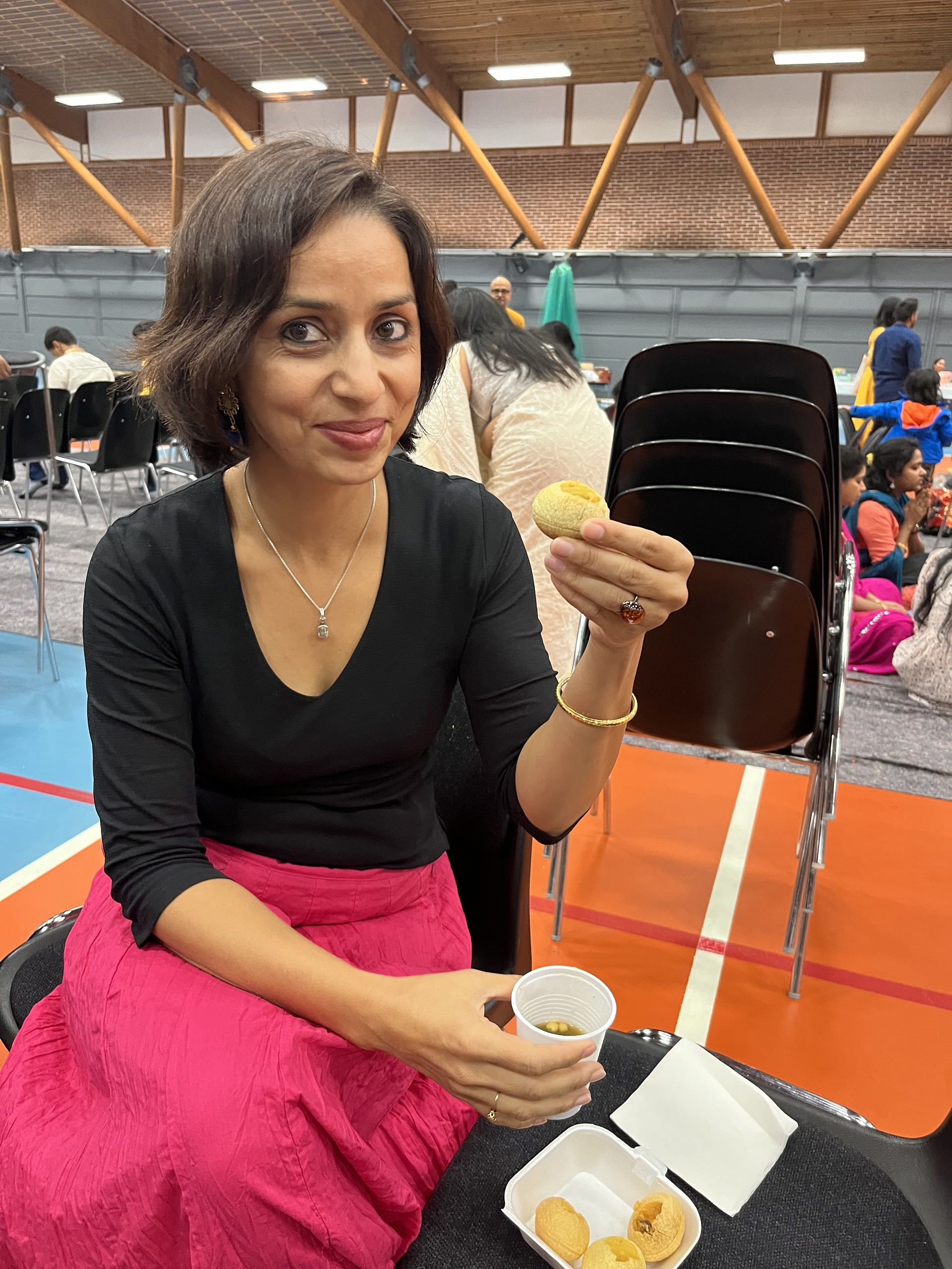 A woman in a black top and pink skirt smiles as she holds up a pani puri, with a cup of tamarind water in her other hand and a box of puris on her lap.