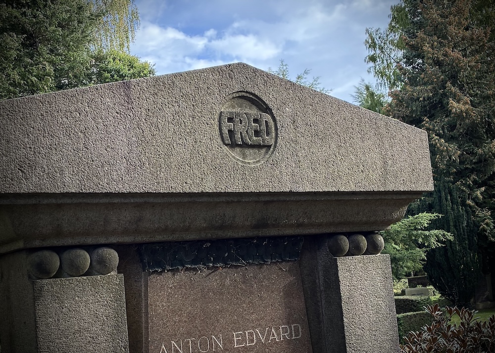 Granite grave monument with FRED carved in a circle at the top and the name Anton Edvard below, surrounded by trees under a cloudy sky