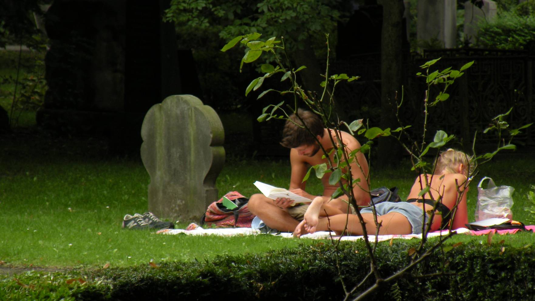 Two people lying on a blanket on the grass in a leafy cemetery, one reading a book, next to an old weathered gravestone