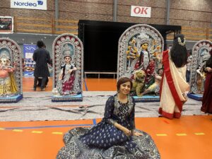 Durga Puja celebration in Copenhagen A smiling woman in a navy and silver Indian dress sits on the floor in front of tall Durga Puja idols of Goddess Durga, Ganesha and other deities, set up in a sports hall.