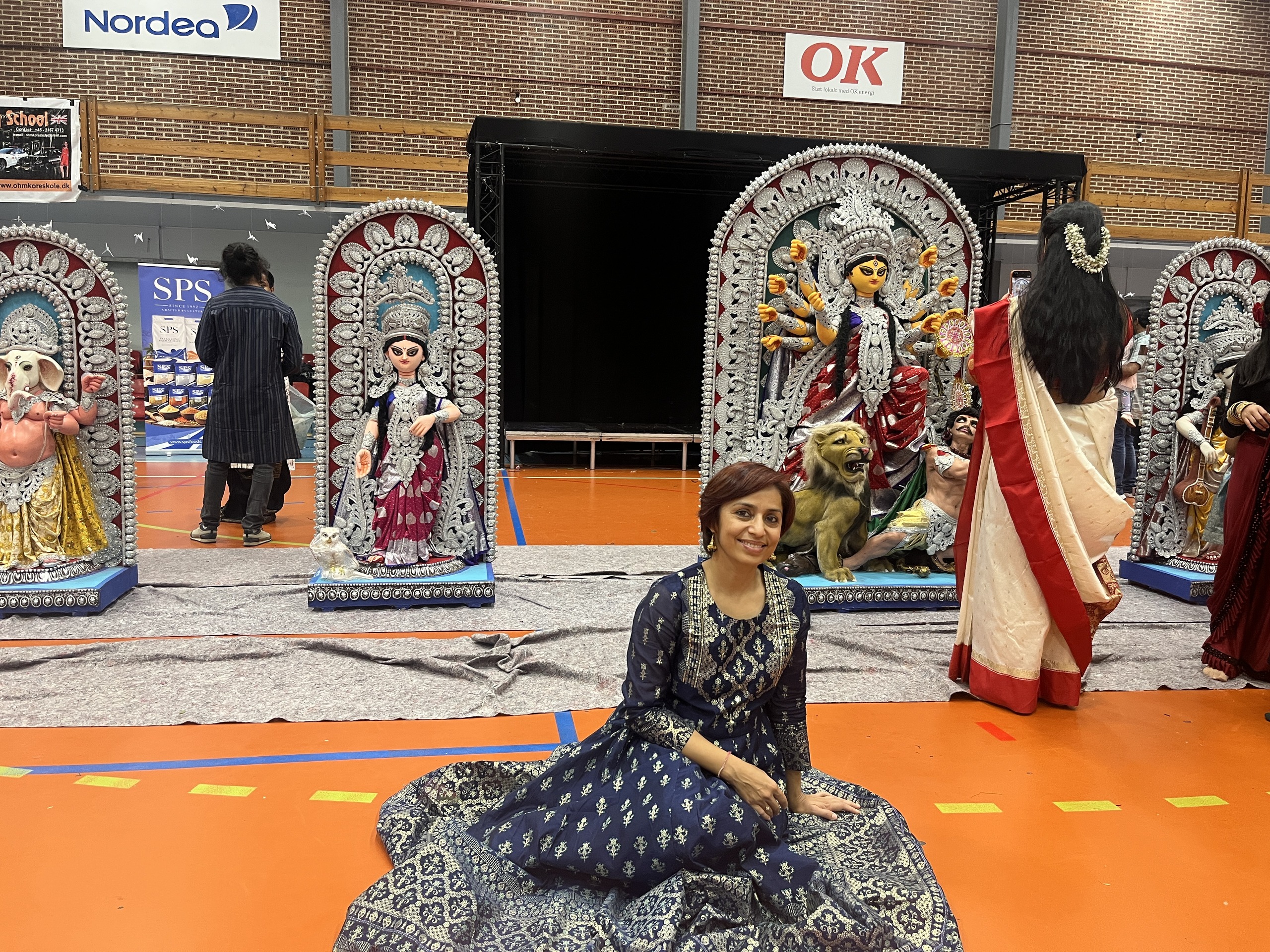 A smiling woman in a navy and silver Indian dress sits on the floor in front of tall Durga Puja idols of Goddess Durga, Ganesha and other deities, set up in a sports hall.